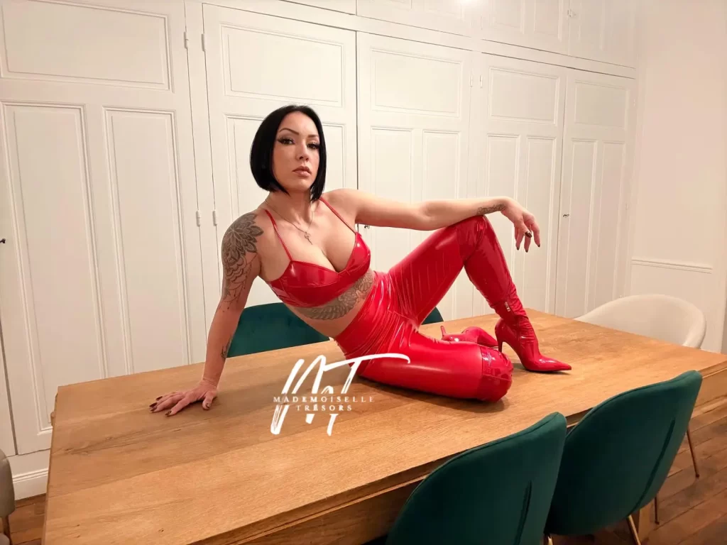 Mademoiselle Trésors. Femme dominatrice portant une tenue en vinyle rouge, un soutien-gorge et un pantalon, posant sur une table en bois devant des chaises vertes et des portes blanches.
